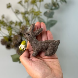 Può includere: Un piccolo coniglio di feltro marrone che tiene un fiore di feltro verde e bianco. Il coniglio è alto circa 7,5 cm ed è un ornamento fatto a mano.