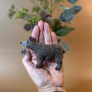 Könnte beinhalten: Handgefertigter Bärenschmuck aus Filz in Braun- und Grautönen, mit detailliertem Gesicht und kleinem, baumelndem Schneeflocken-Anhänger. Der Bär ist etwa 7.6 cm lang, ideal für festliche Dekorationen oder als einzigartiges Geschenk.