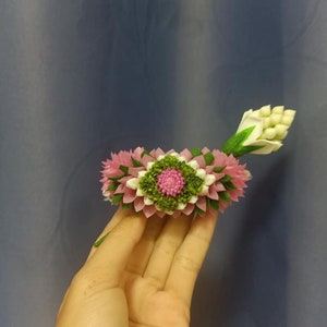 May include: A pink and white flower garland with green leaves, made of fabric, is held in a hand.