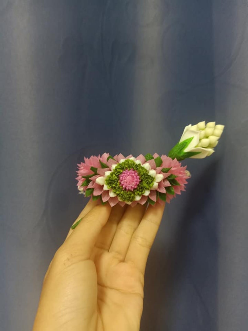 May include: A pink and white flower garland with green leaves, made of fabric, is held in a hand.