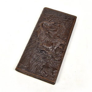 Puede incluir: Cartera de cuero marrón con un diseño de tigre en relieve. La cartera tiene forma rectangular y alargada, con una superficie texturizada y costuras visibles en los bordes. El tigre está representado en un entorno natural.