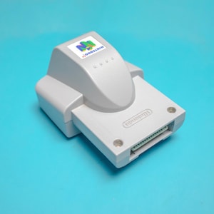 May include: A gray Nintendo 64 Transfer Pak with the Nintendo logo and the words "Transfer Pak" printed on the top. The Transfer Pak is a device that allows players to transfer data between the Nintendo 64 and the Game Boy Color.
