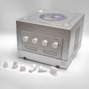 May include: A silver Nintendo GameCube console with four white buttons and a disc slot. The console is sitting on a white surface. There are six white plastic plugs scattered in front of the console.