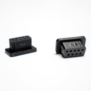 May include: Two black rectangular electronic connectors with multiple holes. One connector is upright, and the other is lying on its side. The connectors are on a white background.