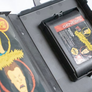 May include: A black Sega Genesis game cartridge with a label featuring the cartoon characters Beavis and Butt-Head. The label is red, white, and black with the text "Beavis and Butt-Head" and "Viacom" printed on it. The cartridge is in a black plastic case.