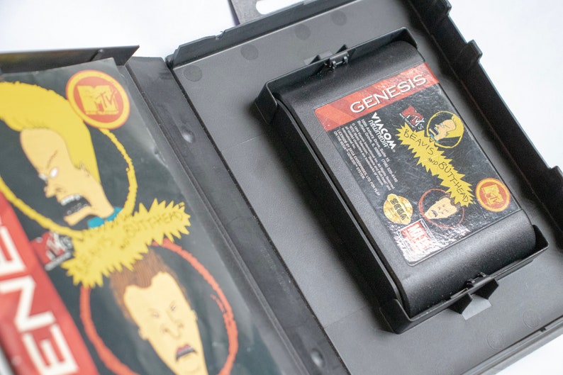 May include: A black Sega Genesis game cartridge with a label featuring the cartoon characters Beavis and Butt-Head. The label is red, white, and black with the text "Beavis and Butt-Head" and "Viacom" printed on it. The cartridge is in a black plastic case.