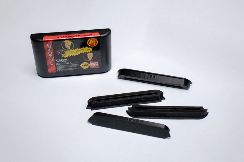 May include: A black Sega Genesis game cartridge for the game "Beavis and Butt-Head" with the text "It's Beavis and Butt-Head" and "VIACOM" printed on the label. Four black plastic cartridge holders are also shown.