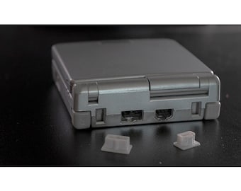 Puede incluir: Una consola de juegos portátil plateada con un diseño rectangular. La consola tiene una tapa con bisagras y varios puertos en la parte inferior. Dos pequeñas cubiertas de botones de color gris claro están delante de la consola. El dispositivo está sobre una superficie oscura.