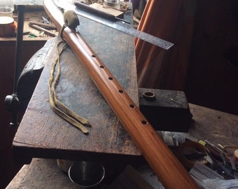 May include: A handcrafted wooden flute with a warm, reddish-brown finish. The instrument features several finger holes and is adorned with a decorative element and leather ties. It rests on a rustic wooden surface.