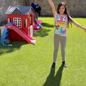 May include: A young girl wearing a pink shirt with the text "Christ with me" in yellow letters, grey pants, and black shoes, is standing on a green lawn with her arms outstretched. She is playing in a backyard with a red and blue plastic playhouse and slide.