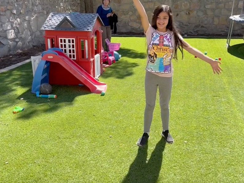 May include: A young girl wearing a pink shirt with the text "Christ with me" in yellow letters, grey pants, and black shoes, is standing on a green lawn with her arms outstretched. She is playing in a backyard with a red and blue plastic playhouse and slide.