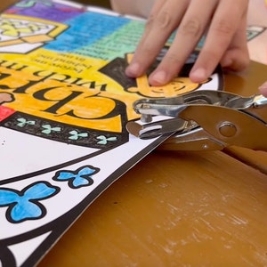 May include: A close-up of a hand using a silver hole punch on a colorful paper with the words "With me, Chris" and a clover design.