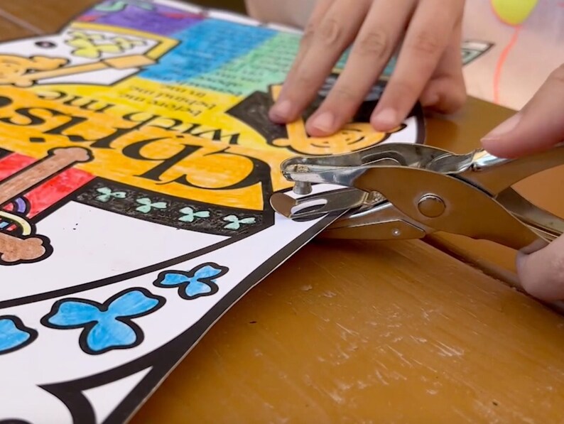 May include: A close-up of a hand using a silver hole punch on a colorful paper with the words "With me, Chris" and a clover design.