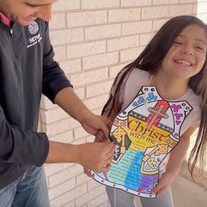 May include: A young girl wearing a colorful paper armor with the words "Christ with me" and a cross. The armor is decorated with flowers and butterflies. A man is helping her put on the armor.