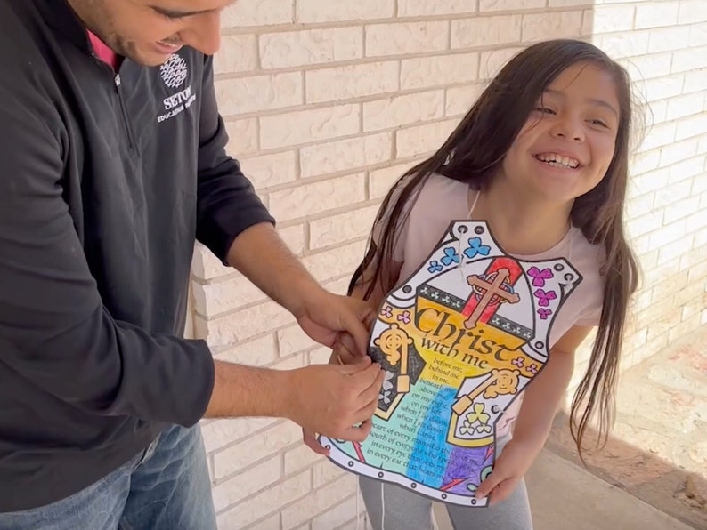 May include: A young girl wearing a colorful paper armor with the words "Christ with me" and a cross. The armor is decorated with flowers and butterflies. A man is helping her put on the armor.