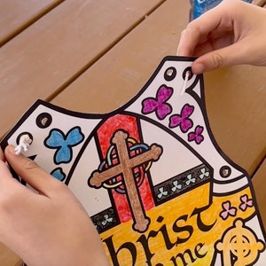 May include: A white paper vest with a colorful design featuring a cross, butterflies, and the text "Christ in me." The vest has holes around the edges for string to be threaded through.