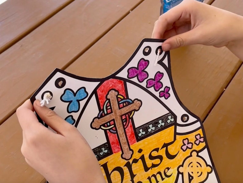 May include: A white paper vest with a colorful design featuring a cross, butterflies, and the text "Christ in me." The vest has holes around the edges for string to be threaded through.