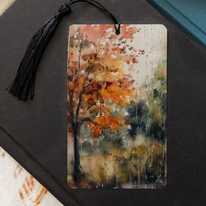 May include: A watercolor bookmark featuring a fall scene with a tree with orange leaves in the rain. The bookmark is rectangular and has a black tassel.