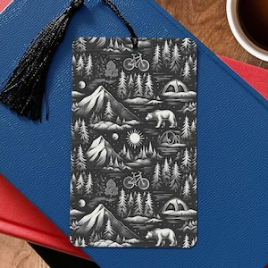May include: Black and white bookmark with a camping and nature theme. The bookmark features a mountain range, trees, a bicycle, a tent, and a bear. The bookmark is made of durable cardstock and has a black tassel.