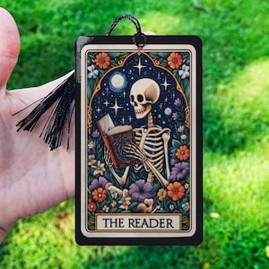 May include: A black bookmark with a colorful illustration of a skeleton reading a book. The bookmark has a black tassel and the text "THE READER" at the bottom.