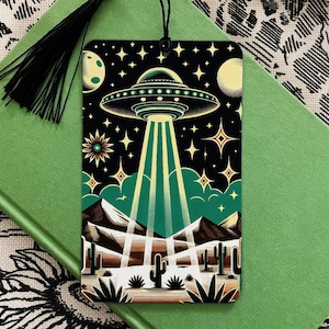 May include: A black and green bookmark featuring a UFO flying over a desert landscape. The UFO is emitting beams of light. The bookmark has a black tassel.