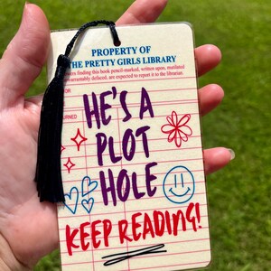 May include: A rectangular bookmark with a cream-colored background featuring red horizontal lines and vertical columns. The text reads PROPERTY OF THE PRETTY GIRLS LIBRARY, HE'S A PLOT HOLE, and KEEP READING! It includes a black tassel and colorful doodles.