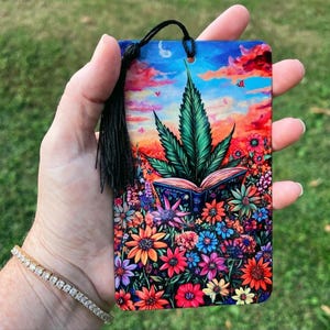May include: A colorful bookmark featuring a green marijuana leaf growing from an open book surrounded by flowers. The background is a sunset sky with clouds and butterflies.