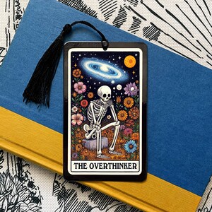 May include: A black bookmark featuring a skeleton seated on a rock amidst a floral arrangement and a galaxy backdrop. The inscription "The Overthinker" is displayed at the base.