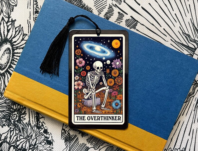 May include: A black bookmark featuring a skeleton seated on a rock amidst a floral arrangement and a galaxy backdrop. The inscription "The Overthinker" is displayed at the base.