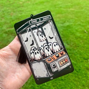 May include: Black rectangular air freshener featuring a claw machine design with white ghost figures and the text "GRAB-A-GHOST". The air freshener has a black tassel and is set against a green background.