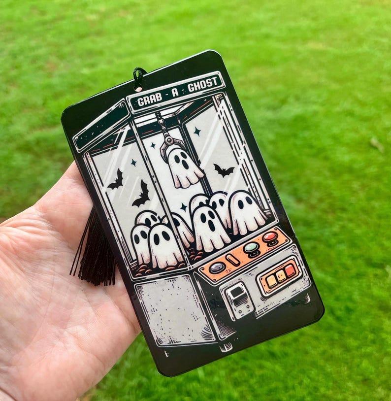 May include: Black rectangular air freshener featuring a claw machine design with white ghost figures and the text "GRAB-A-GHOST". The air freshener has a black tassel and is set against a green background.