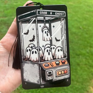 May include: Black rectangular air freshener with a claw machine graphic featuring ghosts and bats. The text "GRAB - A - GHOST" is at the top. A black tassel hangs from the top. The background is green grass.