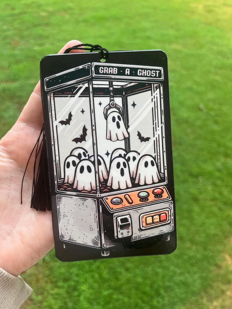 May include: Black rectangular air freshener with a claw machine graphic featuring ghosts and bats. The text "GRAB - A - GHOST" is at the top. A black tassel hangs from the top. The background is green grass.