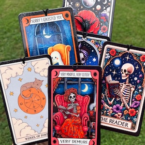 May include: Set of six car air fresheners with colorful illustrations and text. The illustrations include a cat, a skeleton reading a book, a skeleton in a chair, and a moon. The text includes "Sorry I ghosted you", "Wheel of Fortune", "Very Mindful, Very Cutesy", "Very Demure", and "The Reader".