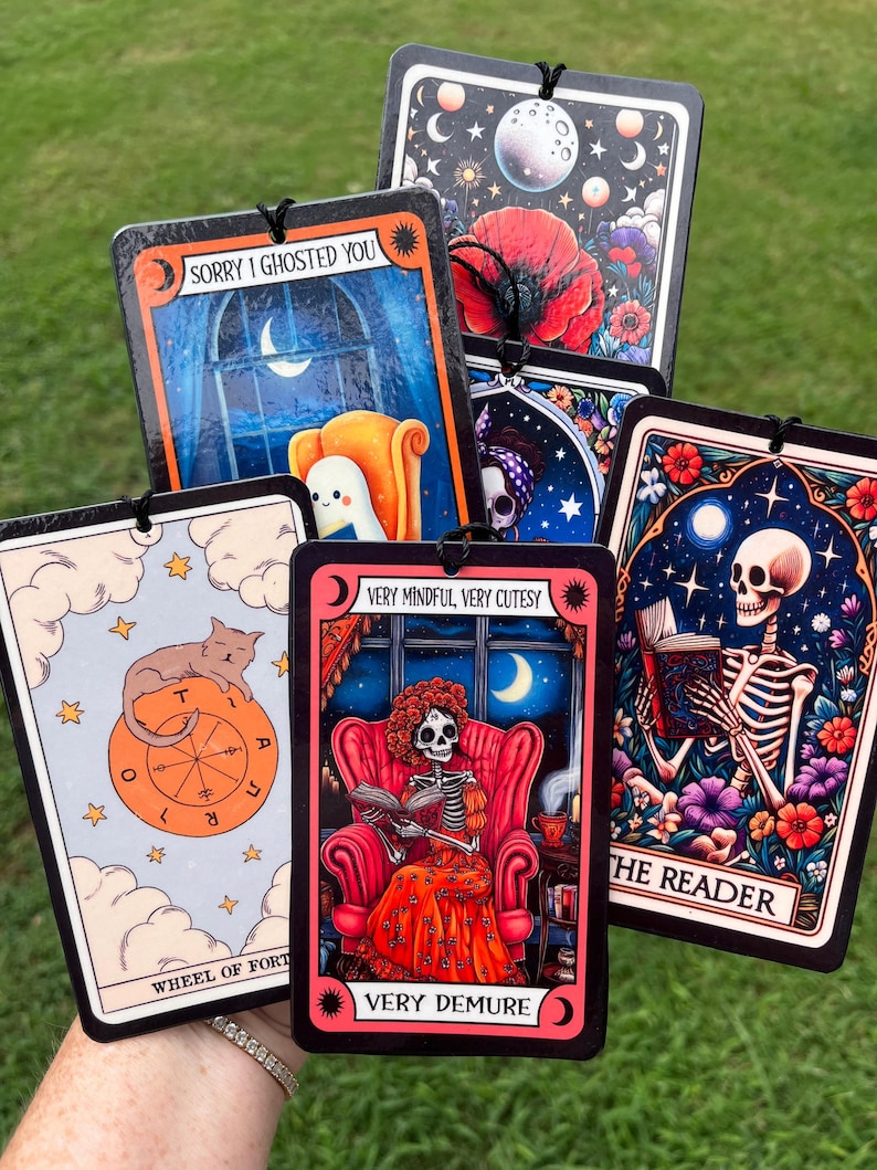 May include: Set of six car air fresheners with colorful illustrations and text. The illustrations include a cat, a skeleton reading a book, a skeleton in a chair, and a moon. The text includes "Sorry I ghosted you", "Wheel of Fortune", "Very Mindful, Very Cutesy", "Very Demure", and "The Reader".