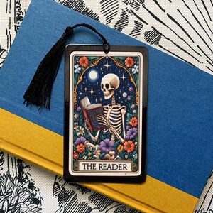 May include: A black bookmark with a colorful illustration of a skeleton reading a book. The bookmark has the text "The Reader" at the bottom.