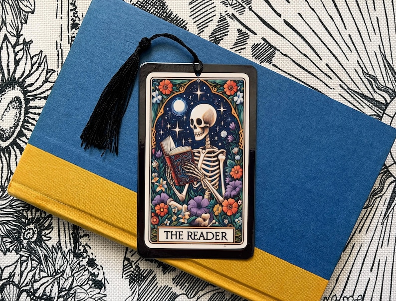 May include: A black bookmark with a colorful illustration of a skeleton reading a book. The bookmark has the text "The Reader" at the bottom.