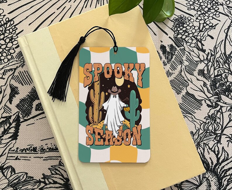 May include: A colorful bookmark with a ghost wearing a cowboy hat and the text "Spooky Season" in a retro font. The bookmark has a tassel and is perfect for Halloween.