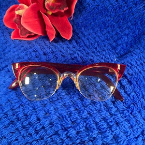 May include: Cat-eye glasses featuring a translucent red upper frame and clear lower lenses. The frames have gold-tone metal accents at the outer corners. A vibrant red orchid flower is positioned above the eyewear.