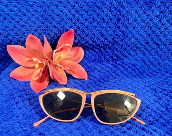 May include: Sunglasses with peach-colored frames and dark lenses, featuring a gold-toned metallic accent along the top brow bar. The sunglasses are positioned next to a pair of artificial peach-colored orchid flowers with green centers.