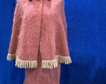 May include: A pink mohair cape with a matching skirt. The cape has a collar, button closure, and fringe trim. The skirt is knee-length and has a similar fringe trim.