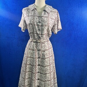 May include: A short-sleeved midi dress featuring a brown and white checkered pattern, a collared neckline, and a full-length button-down front. The garment includes a matching thin belt at the waist and is displayed on a mannequin with a floral hair accessory.