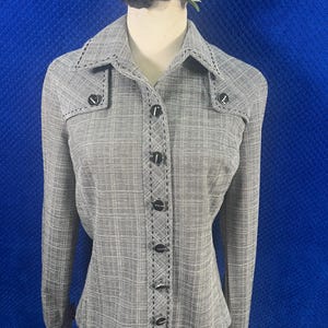 May include: A long-sleeved button-down shirt in a grey and white cross-hatched pattern. The shirt features a pointed collar, decorative black stitching along the placket, collar, and cuffs, and six round black buttons down the front centre.