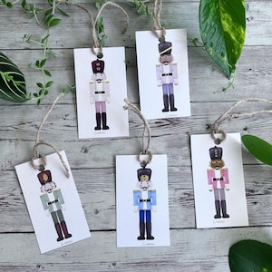May include: Set of five rectangular gift tags featuring watercolor illustrations of nutcracker soldiers in various uniform colors. Each tag is strung with twine and has a small metal eyelet for hanging. The tags are arranged on a wooden surface.