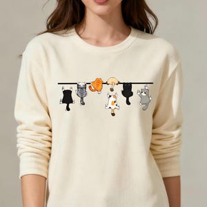 May include: Cream-colored sweatshirt featuring a graphic of six cartoon cats hanging from a black line. The cats are in various colors, including black, gray, orange, and calico. The sweatshirt has long sleeves and a crew neck.