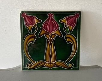 May include: A vintage ceramic tile with a green background and a floral design. The flowers are pink and yellow with intricate details. The tile is square and has a glossy finish.