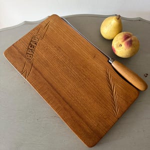 May include: A rectangular wooden bread board with a carved banner containing the word BREAD and a wheat stalk motif. A metal bread knife with a wooden handle is positioned along the side of the board, accompanied by two yellow pears.