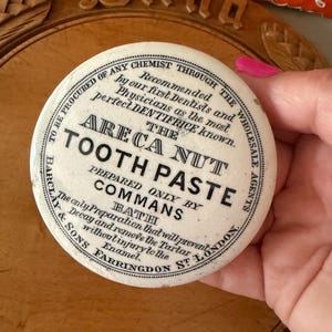 May include: A vintage Areca Nut Tooth Paste container with a round, off-white lid. Black text details the product and manufacturer, "COMMANS BATH LONDON." The container is held by a person. The background includes a wooden breadboard and a decorative orange textile.