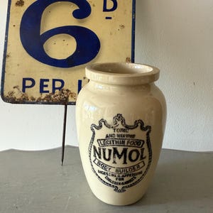May include: A cream-coloured ceramic jar with a blue and black label that reads "TONIC AND NERVINE LECITHIN FOOD NUMOL BODY BUILDER DIGESTIVE & APPETISING FOR CHILDREN & ADULTS".