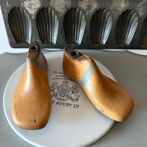 May include: Two vintage wooden shoe lasts with a light brown finish, resting on a white plate with a crest and text. A metal madeleine pan is in the background. The shoe forms have a gray stripe.